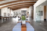 This generously sized single-floor amenity space blends contemporary design with warm, rustic elements to create an inviting and functional gathering environment. The centerpiece of the main lounge is a dramatic floor-to-ceiling stone fireplace, flanked by multiple seating arrangements that encourage relaxation and socializing. Striking curved wooden beams arch across the soaring ceiling, adding architectural character while complementing the polished concrete floors below. Expansive floor-to-ceiling windows and glass doors flood the interior with natural light and provide seamless views of an outdoor pool area and surrounding courtyard. A kitchen or refreshment bar area features granite or quartz countertops, reclaimed wood accents, bar stools, and a large pass-through window overlooking the exterior. A secondary lounge or media room showcases a reclaimed wood accent wall with a wall-mounted television, offering a more intimate retreat. Additional seating nooks with soft furnishings, decorative mirrors, and curated artwork are distributed throughout the space. Reclaimed wood columns and beam accents tie the design together, creating a cohesive rustic-modern aesthetic throughout this well-appointed communal facility.