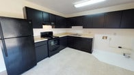 This single-floor space is defined by a clean, functional layout designed to maximize utility and visual cohesion. The kitchen is a standout feature, showcasing bold dark cabinetry that lines the walls in an L-shaped configuration, offering generous storage across both upper and lower cabinets. The rich, near-black finish of the cabinetry creates a striking contrast against the light-toned countertops, giving the kitchen a modern and polished aesthetic. A full suite of matching black appliances — including a refrigerator, electric range with oven, and dishwasher — integrates seamlessly with the cabinetry for a unified look. An under-cabinet range hood is positioned above the stove, providing practical ventilation. The sink is set into the corner of the countertop, making efficient use of the kitchen's layout. Light tile flooring runs throughout, brightening the space and complementing the bold upper elements. Overhead fluorescent lighting ensures the kitchen is well-lit and functional at any time of day. The textured ceiling adds subtle depth to the space. Overall, the kitchen presents a crisp, coordinated environment that balances practicality with a strong design sensibility.