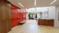 This yoga studio is housed in a thoughtfully designed, single-floor space that makes a strong first impression through its contemporary lobby. The entry area is defined by a dramatic, high-gloss red accent wall that commands attention and energizes the space, complemented by light-toned large-format tile flooring that extends throughout. Sleek linear recessed lighting runs along the ceiling, creating a clean, modern aesthetic while ensuring the space is well-lit at all times.

Large glass doors at the entrance allow natural light to pour in, offering views of the surrounding urban streetscape and fostering a sense of openness and connection to the outdoors. A reception desk finished in warm wood tones provides a welcoming check-in point for visitors, while a minimalist bench along the red wall offers convenient seating near the entrance.

Curated artwork adorns the walls throughout, contributing to a calm yet energetic atmosphere befitting a wellness environment. Lush potted plants and vibrant floral arrangements add organic warmth and vitality to the space. An elevator provides access and convenience, and the corridor area features additional framed artwork and sculptural wall installations, reinforcing the studio's commitment to a refined, inspiring atmosphere. The overall design balances bold visual elements with a serene ambiance ideal for a yoga practice.