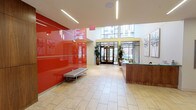 This yoga studio is housed in a thoughtfully designed, single-floor space that makes a strong first impression through its contemporary lobby. The entry area is defined by a dramatic, high-gloss red accent wall that commands attention and energizes the space, complemented by light-toned large-format tile flooring that extends throughout. Sleek linear recessed lighting runs along the ceiling, creating a clean, modern aesthetic while ensuring the space is well-lit at all times.

Large glass doors at the entrance allow natural light to pour in, offering views of the surrounding urban streetscape and fostering a sense of openness and connection to the outdoors. A reception desk finished in warm wood tones provides a welcoming check-in point for visitors, while a minimalist bench along the red wall offers convenient seating near the entrance.

Curated artwork adorns the walls throughout, contributing to a calm yet energetic atmosphere befitting a wellness environment. Lush potted plants and vibrant floral arrangements add organic warmth and vitality to the space. An elevator provides access and convenience, and the corridor area features additional framed artwork and sculptural wall installations, reinforcing the studio's commitment to a refined, inspiring atmosphere. The overall design balances bold visual elements with a serene ambiance ideal for a yoga practice.