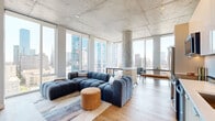 This contemporary high-rise residence masterfully blends industrial and modern design, defined by its signature exposed concrete ceilings and structural columns that lend a striking architectural character throughout. The open-concept living area is a standout, wrapped in expansive floor-to-ceiling windows on multiple sides that flood the space with natural light and frame panoramic city skyline views. Light-toned hardwood flooring runs continuously throughout, enhancing the sense of openness and warmth.

The kitchen is outfitted with a full suite of stainless steel appliances, including a French door refrigerator, range, and dishwasher, complemented by clean white cabinetry, a subway tile backsplash, and light wood accent panels for a refined, contemporary aesthetic.

The bedrooms offer a calm retreat, each featuring the same exposed concrete ceiling detail, hardwood floors, and generous windows with city views. The primary suite connects to a well-appointed bathroom featuring a long vanity with warm wood cabinetry, white quartz countertops, and illuminated mirrors, with a walk-in closet adjacent. Additional bathrooms are clean and functional with light tile flooring. A hallway with artwork display space connects the private areas, completing this thoughtfully designed urban residence.