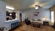 This single-floor residence features an open-concept design that integrates the kitchen and living area into one cohesive, functional space. The living room is generously proportioned, anchored by a ceiling fan with a light fixture that provides both airflow and ambient lighting. Rich hardwood-style flooring runs throughout, complemented by a large, ornate area rug in warm red and gold tones that defines the seating area. The living space is furnished with a cream sofa adorned with decorative pillows in blue, yellow, and patterned accents, paired with a classic leather armchair and a dark wood coffee table. A colorful abstract painting adds a focal point of visual interest to the neutral gray walls. The kitchen features dark cabinetry contrasted by a light stone countertop, with bar-height seating along the peninsula offering a casual dining option. A pendant light illuminates the counter workspace. A doorway visible from the main living area leads to additional rooms, suggesting a practical and well-connected floor plan. The overall ambiance is warm, inviting, and well-appointed, balancing comfort with a stylish, contemporary aesthetic.