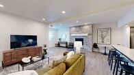 Located at 1900 Poplar St in Forest Grove, Oregon, this single-floor community amenity center offers an impressive array of shared spaces thoughtfully designed for recreation and socializing. The combined kitchen and living room serves as the heart of the facility, featuring a modern kitchen with rich dark cabinetry, light quartz countertops, stainless steel appliances, a tiled backsplash, and a breakfast bar with seating. The adjacent living area is anchored by a striking stacked-stone fireplace and includes comfortable lounge seating, a large flat-screen TV, and light hardwood-style flooring throughout. A connecting hallway leads to the remaining amenity spaces, including the game room and a well-equipped exercise room. The fitness space features durable rubberized flooring, abundant natural light from large windows overlooking the greenery outside, and free weight equipment. The highlight of the facility is the expansive indoor sport court with a bright blue and green surface suitable for pickleball and similar activities, topped with a translucent roof that floods the space with daylight. Overhead roll-up doors open the court directly to the outdoor patio area, which includes a stone-clad outdoor grill station, picnic-style seating, and a large wooden playground structure — creating a seamless indoor-outdoor experience ideal for community gatherings.