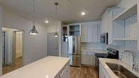 Located at 1817 Sir Tyler Dr in Wilmington, NC, this single-story space showcases a thoughtfully designed interior with a strong emphasis on the kitchen as a central feature. The kitchen is outfitted with crisp white shaker-style cabinetry that extends to the ceiling, providing generous storage capacity. Stainless steel appliances, including a French door refrigerator, microwave, and range, lend a sleek, contemporary feel to the space. A large white quartz island anchors the kitchen, offering ample prep and gathering space. Hexagonal white tile backsplash adds a subtle decorative touch against the clean cabinetry. Warm wood-tone flooring runs throughout, providing a natural contrast to the bright, neutral palette. Recessed lighting combined with two brushed nickel pendant lights hanging above the island ensure the space is well-lit and inviting. Built-in open shelving near the refrigerator offers additional display or storage options. The overall ambiance is bright and modern, with a clean aesthetic that balances functional design with contemporary style.
