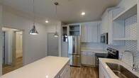 Located at 1817 Sir Tyler Dr in Wilmington, NC, this single-story space showcases a thoughtfully designed interior with a strong emphasis on the kitchen as a central feature. The kitchen is outfitted with crisp white shaker-style cabinetry that extends to the ceiling, providing generous storage capacity. Stainless steel appliances, including a French door refrigerator, microwave, and range, lend a sleek, contemporary feel to the space. A large white quartz island anchors the kitchen, offering ample prep and gathering space. Hexagonal white tile backsplash adds a subtle decorative touch against the clean cabinetry. Warm wood-tone flooring runs throughout, providing a natural contrast to the bright, neutral palette. Recessed lighting combined with two brushed nickel pendant lights hanging above the island ensure the space is well-lit and inviting. Built-in open shelving near the refrigerator offers additional display or storage options. The overall ambiance is bright and modern, with a clean aesthetic that balances functional design with contemporary style.
