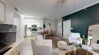 Located in Grand Prairie, Texas, this 1-bedroom, 1-bathroom residence offers a thoughtfully designed open-concept layout that seamlessly connects the living, dining, and kitchen areas. The living room is a standout space, anchored by a bold deep teal accent wall, crown molding, a ceiling fan, and large sliding glass doors fitted with vertical blinds that invite abundant natural light. The kitchen is outfitted with white shaker-style cabinetry, granite countertops, stainless steel appliances, and a peninsula that provides additional seating and workspace. The adjacent dining area features hardwood-style flooring, crown molding, and a round gold-framed mirror that adds a decorative focal point. The bathroom showcases a wide granite-topped vanity with white cabinetry, a large wood-framed mirror, and modern light fixtures. The bedroom includes a walk-in closet with built-in shelving and carpeted flooring, offering practical storage solutions. A dedicated laundry area adds everyday convenience. Throughout the home, consistent hardwood-style flooring, neutral tones, and clean architectural lines create a cohesive and modern aesthetic well-suited for comfortable living.