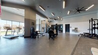 This contemporary fitness center is thoughtfully designed to support a full range of workout routines. The main exercise floor is expansive and open, finished with durable speckled rubber flooring that provides both safety and comfort underfoot. Sleek linear LED lighting runs across the gray-toned ceiling, complemented by large ceiling fans that maintain comfortable airflow throughout the space.

The strength training zone features a comprehensive selection of cable and resistance machines, a multi-function power rack, and a full dumbbell rack displayed along a mirrored wall. Mounted televisions are positioned to keep users engaged during their sessions. A separate cardio area houses a row of treadmills positioned facing large exterior windows, offering views to the outdoors while exercising.

A dedicated studio space is enclosed with floor-to-ceiling glass walls and doors, providing a versatile area for cycling, group fitness, or individual training sessions. Cardio equipment including stair climbers and stationary bikes are also available in adjacent zones.

A utility area near the entry provides neatly stacked towels in vibrant colors, alongside a linen collection bin — ensuring convenience and cleanliness for all users. The overall design is modern and functional, with a neutral gray and white color palette that gives the facility a clean, professional atmosphere.