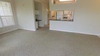This single-floor residence offers a clean, open-concept layout that connects the living area and kitchen in a cohesive, functional flow. The spacious main living area is finished with wall-to-wall plush carpeting in a warm neutral tone, creating a comfortable and inviting atmosphere. Soft, light-colored walls reflect the available light, contributing to an airy and open feel throughout the space.

The kitchen is thoughtfully designed with a peninsula-style breakfast bar that separates it from the living area while maintaining visual connectivity. Dark countertops provide a subtle contrast against the white cabinetry, and white appliances — including a full-size refrigerator and range — are neatly integrated into the layout. A secondary counter area is visible along the adjacent wall, offering additional workspace and storage.

A hallway leads further into the residence, hinting at additional rooms beyond the main living space, while a doorway adds to the practical flow of the floor plan. The overall design emphasizes simplicity and functionality, with a neutral palette throughout that lends itself well to a variety of interior styles. The layout is well-suited for everyday living, offering a welcoming and organized environment from the moment you step inside.