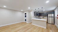 This single-floor space showcases a clean, modern design with an open-concept layout that seamlessly connects the living area to the kitchen. Light-toned hardwood flooring flows throughout, creating a bright and cohesive aesthetic complemented by recessed lighting overhead.

The kitchen is a standout feature, equipped with stainless steel appliances including a range, over-the-range microwave, dishwasher, and refrigerator. Two-toned cabinetry pairs charcoal upper cabinets with warm natural wood lower cabinets, accented by gold hardware. A dramatic marble-look backsplash with bold veining adds visual depth, while the white quartz peninsula countertop provides additional prep space and serves as a natural divider between the kitchen and living area. Sleek black pendant lights with gold accents hang above.

The open living area is generously sized, offering flexible use of space, with a wall-mounted AC unit and baseboard heating ensuring year-round comfort. Natural light enters through windows fitted with white blinds.

A dedicated laundry nook houses a stacked washer and dryer unit, conveniently accessible from the main living space. The bathroom features clean white tile flooring, a vanity with understated cabinetry, and brushed nickel fixtures, completing this well-appointed and functional space.