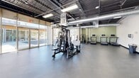 Located at 611 S Thornton Ave in Dalton, Georgia, this single-floor fitness facility offers a generously sized, open-plan workout environment designed to accommodate a comprehensive range of exercise activities. The space features an industrial-modern aesthetic, with high ceilings showcasing exposed steel trusses and large silver HVAC ductwork that runs throughout, contributing to an airy, warehouse-style ambiance.

Abundant natural light pours in through expansive floor-to-ceiling glass walls and large storefront windows along the perimeter, providing clear views of the surrounding streetscape and keeping the interior bright and welcoming. The durable, rubber-coated flooring spans the main workout area, offering both safety and functionality underfoot.

The facility is well-equipped with a variety of strength and cardio machines, including multi-station cable systems, treadmills, stationary bikes, rowing machines, and free weights. A dedicated area features stability balls, medicine balls, and benches for functional training. White-painted brick columns add architectural character throughout the space, while glass-paneled interior doors lead to additional areas such as restrooms. Bright overhead fluorescent lighting supplements the natural light, ensuring a well-illuminated environment throughout the day. The overall layout is open and versatile, supporting individual workouts and group training alike.
