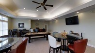 Located at 301 Barcelona Dr in Fayetteville, NC, this single-floor community space is designed for social gathering and recreation. The main room features a tray ceiling with recessed lighting and a ceiling fan, lending the space a polished, well-appointed feel. A full-size pool table serves as the centerpiece of the room, complemented by a wall-mounted flat-screen television ideal for group viewing. Plush carpeting adds warmth underfoot, while neutral wall tones create a clean, welcoming atmosphere. A round high-top dining table with mixed seating — including leather and upholstered chairs — offers a casual spot for conversation or games. Toward the rear of the space, a dedicated lounge area with comfortable seating provides a relaxed retreat, accented by decorative elements including a large wall clock. A workstation area along one wall adds a functional touch for residents needing a quiet place to focus. Natural light filters through the space, enhanced by the layered lighting system embedded in the tray ceiling. The overall design balances leisure and comfort, making it a versatile and inviting destination for community members to gather, unwind, and connect.