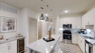 Located in Dallas, TX, this single-story space showcases a contemporary design with a bright, neutral palette throughout. The kitchen serves as a central highlight, featuring a large island topped with speckled granite countertops that provide both functionality and visual appeal. Three metallic pendant lights hang above the island, adding an elegant and modern touch to the space.

The kitchen is outfitted with stainless steel appliances, including a gas range and an over-the-range microwave, complemented by crisp white cabinetry that extends to the upper walls. A subtle geometric tile backsplash adds texture behind the cooking area, while recessed lighting ensures the space remains bright and well-illuminated. A built-in wine cooler is tucked neatly beneath the counter, offering a refined amenity for entertaining.

The light-toned flooring flows seamlessly throughout, contributing to an open and airy atmosphere. Clean white walls and smooth ceilings enhance the sense of space, while interior doors maintain the cohesive, minimalist aesthetic. Overall, the design balances practicality with modern style in a well-appointed Dallas residence.