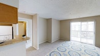 This single-story space offers a straightforward, functional layout designed for comfortable everyday living. The main living area is finished with wall-to-wall carpeting in a warm, neutral tone that complements the beige walls throughout. A textured popcorn ceiling adds a classic touch overhead, while clean white baseboards provide a crisp contrast along the perimeter of the room.

Natural light enters generously through a sliding glass door positioned along the exterior wall, which opens to a private balcony enclosed by a railing — an ideal spot for fresh air and outdoor views. Vertical blinds offer light control and privacy as needed.

Adjacent to the living area, a partial view into a hallway reveals access to additional rooms within the unit, suggesting a well-connected interior flow. A countertop with a tile or stone finish is visible near what appears to be a kitchen transition area, hinting at a practical open connection between living and kitchen spaces. A small decorative plant adds a touch of life to this counter area.

Overall, the space presents a clean, neutral palette and a comfortable, open atmosphere suited for a variety of furnishing arrangements.