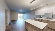 This well-appointed one-bedroom, one-bathroom unit offers a thoughtfully designed open-concept layout across a single floor. The combined kitchen and living area forms the heart of the unit, showcasing a contemporary aesthetic with wood-look flooring that flows seamlessly throughout the space.

The kitchen is a standout feature, equipped with two-tone cabinetry pairing white upper cabinets with warm wood-finish lower cabinets, a mirrored backsplash, white quartz countertops, and a full suite of stainless steel appliances including a refrigerator, range with hood, and dishwasher. A central island with pendant lighting above provides additional workspace and a casual dining option.

The living area extends toward a full-width sliding glass door that opens onto a private balcony, bringing in natural light and outdoor views. The bedroom is a comfortable size, featuring the same wood-look flooring and its own sliding glass door access to the balcony.

The bathroom is finished with a wood-tone vanity, white countertop, and a large mirror. A dedicated laundry room houses a stacked washer and dryer unit alongside the water heater, adding practical convenience. The unit also includes a closet for additional storage.