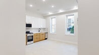 Located at 40 Lembeck Ave in Jersey City, NJ, this thoughtfully designed single-floor unit makes efficient use of its intimate footprint. The open-concept kitchen is a standout feature, showcasing a contemporary two-tone design with glossy white upper cabinetry and warm natural wood lower cabinets. A full suite of stainless steel appliances — including a gas range, refrigerator, built-in microwave, and dishwasher — is complemented by a vertical tile backsplash and a sleek undermount sink. Natural light filters in through a window adjacent to the kitchen, enhancing the bright, airy atmosphere created by recessed ceiling lighting throughout. The living area is finished with light-toned flooring, clean white walls, and a wall-mounted mini-split HVAC unit, providing year-round climate control. A window overlooking a greenery-filled outdoor area adds a refreshing touch of nature to the space. The unit includes two bathrooms: one features a soaking tub with elegant vertically-stacked mosaic tile surround, while the other offers a clean, minimalist design with a white vanity and toilet. A hallway with louvered doors provides additional storage or utility access, completing this well-appointed space.