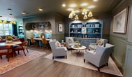 This 2,310-square-foot single-floor amenity space is part of a modern multi-story residential community, offering residents a thoughtfully curated selection of indoor and outdoor facilities. The fitness center is generously sized and carpeted, housing a comprehensive array of strength and cardio equipment, including cable machines, stationary bikes, and free weights, complemented by ceiling fans, wall-mounted TVs, and mirrored walls that enhance the sense of space. Adjacent to the fitness area is a sophisticated lounge featuring rich hardwood flooring, wainscoting, teal-painted built-in bookshelves, and a chandelier, furnished with plush seating arrangements ideal for relaxation or socializing. A separate sitting room offers an intimate setting with wingback chairs, a round coffee table, and curated wall art. The leasing or co-working area includes a central work desk, recessed lighting, and tasteful décor. A powder room features bold botanical wallpaper, geometric-patterned tile flooring, navy wall tile, and antique brass fixtures. Outdoors, landscaped walkways, ornamental plantings, a gated pool area, and a cornhole lawn game area provide residents with inviting spaces for recreation and leisure.