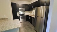 This single-floor space is designed with a contemporary aesthetic that emphasizes clean lines and modern finishes. The kitchen serves as a standout feature, outfitted with a full suite of stainless steel appliances including a refrigerator, electric range, over-the-range microwave, and dishwasher. Dark espresso cabinetry extends from floor to ceiling, providing generous storage while creating a bold visual contrast against the light granite countertops. A neutral tile backsplash adds texture and depth to the space, complementing the overall color palette. The sink is positioned beneath upper cabinets along the back wall, making efficient use of the kitchen's L-shaped layout. A peninsula counter extends outward, offering additional prep space and the potential for casual dining or bar seating. The flooring throughout is a warm-toned hardwood-style plank, lending a grounded, cohesive feel to the interior. The cabinetry hardware and fixtures maintain a consistent modern style, tying all elements together. Overall, this space delivers a functional yet refined environment well-suited for everyday living, with thoughtful design choices that balance practicality and contemporary style.