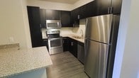 This single-floor space is designed with a contemporary aesthetic that emphasizes clean lines and modern finishes. The kitchen serves as a standout feature, outfitted with a full suite of stainless steel appliances including a refrigerator, electric range, over-the-range microwave, and dishwasher. Dark espresso cabinetry extends from floor to ceiling, providing generous storage while creating a bold visual contrast against the light granite countertops. A neutral tile backsplash adds texture and depth to the space, complementing the overall color palette. The sink is positioned beneath upper cabinets along the back wall, making efficient use of the kitchen's L-shaped layout. A peninsula counter extends outward, offering additional prep space and the potential for casual dining or bar seating. The flooring throughout is a warm-toned hardwood-style plank, lending a grounded, cohesive feel to the interior. The cabinetry hardware and fixtures maintain a consistent modern style, tying all elements together. Overall, this space delivers a functional yet refined environment well-suited for everyday living, with thoughtful design choices that balance practicality and contemporary style.
