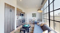 This thoughtfully designed one-bedroom, one-bathroom unit makes efficient use of its compact footprint without sacrificing style or functionality. The open-plan living area is the heart of the space, anchored by dramatic floor-to-ceiling steel-framed windows that flood the room with natural light and frame sweeping urban views. Exposed ductwork at the ceiling adds an industrial character that complements the light hardwood flooring running throughout.

The kitchen is fully equipped with stainless steel appliances, including a full-sized range, refrigerator, and dishwasher, paired with wood-toned cabinetry and a white subway tile backsplash. A small dining area sits adjacent, offering a dedicated space for meals. A tall storage cabinet provides additional organization within the living zone.

The bedroom is a private, well-proportioned retreat featuring a high clerestory window that introduces natural light while maintaining privacy. Clean white walls and recessed lighting contribute to a calm, restful atmosphere.

The bathroom is finished with white subway tile in a glass-enclosed walk-in shower with a linear drain and brushed nickel fixtures, reflecting a clean, contemporary aesthetic. A hallway connects all areas of the unit efficiently, completing a practical and polished layout.