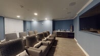 Located at 25500 Westheimer Pkwy in Katy, TX, this dedicated home theater room delivers a true cinema experience within a single-floor layout. The space is defined by its cool blue-toned walls accented with crown molding and recessed lighting, creating an immersive and focused atmosphere ideal for screenings.

Multiple rows of plush, patterned recliner-style theater chairs are arranged to face a large motorized projection screen along the front wall, flanked by ambient LED backlighting that adds a cinematic glow. A wall-mounted flat-screen television is also present, offering additional viewing flexibility.

At the rear of the room, a well-appointed wet bar area features dark wood cabinetry with a granite or stone-look countertop, a stainless steel mini-refrigerator, a built-in microwave, and a dedicated wine rack — providing everything needed for convenient in-room refreshments. A media storage rack is also visible, keeping physical media neatly organized.

The flooring transitions between hardwood-style planks and carpet underfoot near the seating area, enhancing comfort during extended viewing sessions. Ceiling-mounted in-wall speakers support a surround sound setup. The room is accessed through a classic white paneled door and is thoughtfully designed to balance both function and comfort for an upscale entertainment experience.