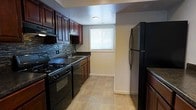 This single-floor residence offers a thoughtfully designed living space with a functional and attractive layout. The kitchen stands out as a central feature, showcasing rich dark-wood cabinetry that provides generous storage alongside a sleek, coordinated aesthetic. A decorative glass tile backsplash adds a modern touch, complementing the dark countertops and lending the space a polished, contemporary feel. Black appliances, including a gas range, over-the-range microwave, and a full-size refrigerator, are seamlessly integrated into the kitchen design. The sink is positioned beneath a window that allows natural light to brighten the space. Light-toned ceramic tile flooring runs throughout, offering a clean contrast to the darker cabinetry and appliances while also providing durability and ease of maintenance. The neutral wall tones create a versatile backdrop that complements the overall design. The kitchen layout is efficiently organized, maximizing workspace and storage without sacrificing flow or accessibility. The overall ambiance of the space is clean, modern, and move-in ready, combining practical amenities with tasteful design elements that make for a comfortable and attractive living environment.