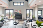 Located at 5500 Riverwood Dr in Flint, Michigan, this two-floor community facility offers a thoughtfully designed layout across a generous footprint. The main floor serves as the heart of the space, anchored by a dramatic great room boasting soaring vaulted ceilings with a striking dark-paneled peak, clerestory windows that flood the area with natural light, and a sleek fireplace framed by French doors that open to an outdoor deck. Wide-plank wood-look flooring flows throughout, complementing the modern, neutral color palette. Adjacent to the great room, a spacious kitchen impresses with white shaker cabinetry, dark granite countertops, a mosaic tile backsplash, a center island, stainless steel appliances, and an elegant chandelier overhead. The dining room provides a comfortable gathering area with round tables, blue upholstered seating, and built-in cabinetry for storage and display. A welcoming entrance with stone tile flooring and a den suitable for meetings or leasing activities round out the main level. The lower floor houses a well-equipped exercise room featuring cardio machines, strength equipment, wall mirrors, and resilient flooring, catering to an active lifestyle. Two staircases connect the floors, ensuring easy circulation throughout this versatile and inviting community space.