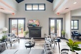 Located at 5500 Riverwood Dr in Flint, Michigan, this two-floor community facility offers a thoughtfully designed layout across a generous footprint. The main floor serves as the heart of the space, anchored by a dramatic great room boasting soaring vaulted ceilings with a striking dark-paneled peak, clerestory windows that flood the area with natural light, and a sleek fireplace framed by French doors that open to an outdoor deck. Wide-plank wood-look flooring flows throughout, complementing the modern, neutral color palette. Adjacent to the great room, a spacious kitchen impresses with white shaker cabinetry, dark granite countertops, a mosaic tile backsplash, a center island, stainless steel appliances, and an elegant chandelier overhead. The dining room provides a comfortable gathering area with round tables, blue upholstered seating, and built-in cabinetry for storage and display. A welcoming entrance with stone tile flooring and a den suitable for meetings or leasing activities round out the main level. The lower floor houses a well-equipped exercise room featuring cardio machines, strength equipment, wall mirrors, and resilient flooring, catering to an active lifestyle. Two staircases connect the floors, ensuring easy circulation throughout this versatile and inviting community space.