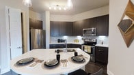 This single-floor residence showcases a thoughtfully designed open-concept layout centered around a striking modern kitchen. The kitchen is appointed with rich, dark espresso-toned cabinetry that extends to the upper walls, providing generous storage while creating a bold visual contrast against the crisp white countertops. Under-cabinet lighting illuminates the classic subway tile backsplash, adding both functionality and a polished aesthetic to the space.

A full suite of stainless steel appliances — including a French door refrigerator, electric range, and built-in microwave — lends the kitchen a professional, contemporary feel. The expansive white island serves as the heart of the kitchen, offering a built-in sink, additional prep space, and casual dining seating, making it ideal for both everyday meals and entertaining guests.

A stylish multi-bulb pendant light fixture anchors the island area, complementing the track lighting overhead that ensures the space is well-lit throughout. The dark hardwood-style flooring grounds the room and ties together the overall design palette. The combination of dark cabinetry, bright countertops, and stainless finishes creates a sophisticated, high-contrast aesthetic that is both functional and visually compelling throughout this well-appointed home.