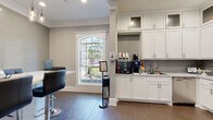 This thoughtfully designed single-floor amenity space blends functionality with refined aesthetics throughout its interconnected areas. The kitchen is a standout feature, outfitted with floor-to-ceiling white shaker cabinetry, a gray mosaic tile backsplash, stainless steel appliances including a side-by-side refrigerator with ice maker, a built-in microwave, dishwasher, and a dedicated coffee and beverage station — all set atop large-format tile flooring. Natural light filters in through an arched, multi-pane window, enhancing the bright and clean atmosphere. Adjacent to the kitchen, a comfortable lounge area welcomes visitors with a pair of patterned accent chairs, a small side table, and a classic wall clock, framed by crisp white wainscoting and soft gray walls. This seating area opens directly to a well-equipped fitness center, visible through wide doorways, featuring cardio machines and ample workout space. A separate media and dining lounge offers a high-gloss mosaic accent wall, a wall-mounted flat-screen television, pendant lighting, and a long bar-height table surrounded by modern black leather swivel stools — ideal for group gatherings. Warm hardwood-style flooring and crown molding tie the spaces together, creating a cohesive and polished environment throughout.