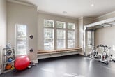 Located at 9125 Highway 6 N in Houston, TX, this fitness center offers a generously sized, single-floor exercise space designed to support a comprehensive workout experience. The room is finished with elegant architectural details, including crown molding and neutral-toned walls, striking a balance between functionality and refined design.

The facility is bathed in abundant natural light, courtesy of large windows lining multiple walls and offering views of the surrounding greenery outdoors. The dark, durable rubber flooring throughout provides both safety and practicality for high-intensity training.

The exercise room is outfitted with a wide array of professional-grade equipment. Cardio machines include treadmills, ellipticals, and cycling bikes, while strength training is well-supported by a Hoist cable machine system, a weight rack with plates, and additional resistance machines. A functional training rig with overhead bars serves as a central feature, ideal for bodyweight and suspension exercises. A stability ball, weight storage rack, and other accessories round out the available equipment.

A floor-to-ceiling mirror along one wall visually expands the space and supports form-checking during workouts. A built-in bench seat beneath the windows adds a thoughtful resting area. Recessed lighting complements the natural light, ensuring the space remains well-lit at all hours.
