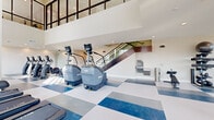 Located at 621 S Sierra Madre St in Colorado Springs, CO, this two-floor fitness facility offers a well-appointed and generously sized workout environment spread across two levels. The ground floor serves as the main exercise area, featuring soaring double-height ceilings with a dramatic open mezzanine above, floor-to-ceiling windows that flood the space with natural light, and sweeping views of the surrounding Colorado landscape and mountain skyline. The distinctive geometric tile flooring in shades of blue, white, and gray adds a dynamic visual element throughout. This level is equipped with a full array of cardio machines — including treadmills, stair climbers, and ellipticals — as well as strength training stations, free weights, and a built-in water station. A sleek staircase with glass and metal railings connects to the upper floor. The second floor houses an additional exercise room finished with light hardwood-style flooring, arched windows framing panoramic mountain views, and a wall-mounted display screen — ideal for group fitness or yoga sessions. A connected living room area offers a more relaxed space. Throughout both levels, the modern architectural design, recessed lighting, clean neutral walls, and glass partitions create a bright, open, and professional fitness environment.