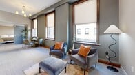 Located on Euclid Avenue in the heart of Cleveland, this two-bedroom, one-bathroom residence offers a thoughtfully designed single-floor layout that maximizes every square foot. The open-concept kitchen and living area serves as the central hub of the home, featuring a sleek kitchen island with dark quartz countertops, white shaker cabinetry extending to the ceiling, a textured tile backsplash, and a full suite of stainless steel appliances. Pendant lighting adds a modern touch above the island. The living area benefits from large, wood-trimmed windows that invite natural light and frame views of the surrounding Cleveland cityscape. The primary bedroom is flanked by dual windows offering elevated urban views, lending an airy quality to the space, while the second bedroom also features a window with natural light and a wall-mounted TV. Both rooms are finished with neutral-toned carpet and warm, understated walls. The bathroom is a standout, showcasing a distinctive star-patterned tile floor, white cabinetry with dark countertops, and a well-lit vanity mirror. A dedicated closet completes the layout. Throughout, the design balances contemporary finishes with classic architectural details, creating a cohesive and functional urban residence.