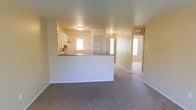 This single-story space features an open-concept design that connects the living area to the kitchen through a half-wall pass-through counter with a dark stone or laminate countertop surface. The kitchen is equipped with white cabinetry and appliances, and benefits from a window that draws in natural light. The living area is finished with wall-to-wall carpeting in a neutral tone, complemented by warm beige walls that create a cohesive, understated palette throughout. Flush-mount ceiling light fixtures provide consistent overhead lighting across the space. A hallway visible beyond the pass-through leads to additional rooms, including what appears to be a room with a built-in shelving unit or closet and a doorway that opens to further spaces with natural light. The layout offers a functional flow between areas, with the open sightlines from the living space to the kitchen enhancing the sense of connectivity. The overall ambiance is clean, neutral, and move-in ready, suited to a variety of furnishing and décor styles.