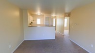 This single-story space features an open-concept design that connects the living area to the kitchen through a half-wall pass-through counter with a dark stone or laminate countertop surface. The kitchen is equipped with white cabinetry and appliances, and benefits from a window that draws in natural light. The living area is finished with wall-to-wall carpeting in a neutral tone, complemented by warm beige walls that create a cohesive, understated palette throughout. Flush-mount ceiling light fixtures provide consistent overhead lighting across the space. A hallway visible beyond the pass-through leads to additional rooms, including what appears to be a room with a built-in shelving unit or closet and a doorway that opens to further spaces with natural light. The layout offers a functional flow between areas, with the open sightlines from the living space to the kitchen enhancing the sense of connectivity. The overall ambiance is clean, neutral, and move-in ready, suited to a variety of furnishing and décor styles.
