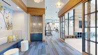 This single-level fitness center is thoughtfully designed with a blend of modern aesthetics and functional layout. Soaring ceilings amplify the sense of openness throughout the space, while expansive floor-to-ceiling windows along multiple walls flood the interior with abundant natural light and provide views of the surrounding outdoor environment. Warm natural wood paneling and trim serve as a unifying architectural element, offering visual warmth against the crisp white walls.

The flooring throughout consists of patterned carpet tiles in varying shades of blue and grey, adding texture and visual interest underfoot. The cardio zone is well-equipped with treadmills, ellipticals, stair climbers, and stationary bikes positioned to take advantage of the outdoor views. A dedicated strength training area houses a cable machine, weight bench, and a full dumbbell rack, accompanied by wall-mounted mirrors that enhance the sense of depth.

A striking accent wall featuring a living green moss installation and geometric wooden paneling anchors the functional training zone, which includes a training rack with resistance bands and medicine balls. The entry vestibule offers built-in blue cubby storage, and the interior is connected by sleek black-framed glass doors, contributing to the space's contemporary, upscale character.
