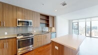 Located in downtown San Diego's vibrant East Village neighborhood, this contemporary one-bedroom, two-bathroom residence offers a well-designed open-concept layout with modern finishes throughout. Warm hardwood flooring flows seamlessly across the entire living space, complementing the clean white walls and creating a bright, inviting atmosphere.

The kitchen and living area share an open, connected layout, anchored by a sleek kitchen featuring flat-front wood-grain cabinetry, a subway tile backsplash, stainless steel appliances including a gas range, built-in microwave, and dishwasher, and a central island with a quartz countertop. Floor-to-ceiling windows and a sliding glass door in the living room flood the space with natural light and frame striking panoramic views of the San Diego skyline.

The bedroom is generously proportioned, with large windows offering additional city views and access to two closets, including a spacious walk-in fitted with multi-tiered shelving and hanging rods. Two full bathrooms serve the unit: one features a glass-enclosed walk-in shower with white tile and an illuminated vanity mirror, while the second offers a full soaking tub and shower combination with similarly clean, contemporary finishes.