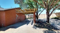 This outdoor car wash facility is a resident-exclusive amenity set within a well-maintained community. The wash area is anchored by a sturdy wood-and-post pergola structure supported by brick-red columns, providing partial shade over a spacious concrete wash bay. The bay features a central floor drain, a mounted pressure hose with spray nozzle, and a token-operated payment kiosk mounted to the adjacent brick wall. A green signage panel clearly displays operating hours and usage requirements, indicating availability from morning through evening for residents only.

The brick building alongside the wash bay is constructed in a style consistent with the surrounding community architecture, featuring a pitched roofline and wall-mounted lighting. The perimeter of the facility is landscaped with decorative gravel beds and mature shade trees, which provide natural canopy and contribute to a clean, organized aesthetic. A privacy fence lines the boundary of the area, creating a defined and enclosed environment.

The broader surroundings include well-kept driveways, green lawns, and multi-story brick residential buildings visible in the background, situating the car wash conveniently within the community layout for easy resident access.
