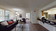 This single-story model unit in Gainesville, Florida presents a practical and well-organized floor plan designed for comfortable everyday living. The living room serves as the heart of the space, featuring wall-to-wall carpet, a textured ceiling, and an exterior door alongside multiple windows that invite natural light and offer views of the surrounding greenery. A wall-mounted television and ample room for seating make it a functional gathering area.

Adjacent to the living room, the kitchen is accessible via a pass-through counter with dark laminate surfaces and bar seating, providing a convenient connection between cooking and living areas. Inside the kitchen, warm wood-toned cabinetry, a stainless steel refrigerator, electric range, and overhead ventilation hood offer a complete cooking setup illuminated by recessed fluorescent lighting.

The unit includes multiple bedrooms, each finished with carpet flooring, a textured ceiling, and windows that bring in natural light. Several bedrooms feature ceiling fans and closet space. The bathrooms are well-appointed, with one showcasing wood-look plank flooring, a vanity with stone-style countertops, a mirror, and a tub-shower combination, while another features a granite-topped vanity and a shower with a curtain enclosure.