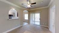 This one-bedroom, one-bathroom single-floor residence offers a well-designed layout that maximizes functionality and comfort. The living area is finished with plush carpeting and features elegant crown molding along the ceiling, adding a refined architectural detail to the space. A ceiling fan with integrated lighting provides both comfort and illumination throughout the room.

Natural light flows generously into the living area through a large sliding glass door fitted with vertical blinds, offering access to an outdoor view framed by surrounding greenery. The arched doorways connecting the living space to the bedroom and other areas lend a classic, welcoming character to the interior design.

The kitchen is accessible via a pass-through counter visible from the living area, creating a semi-open connection between the two spaces while maintaining distinct zones. Neutral-toned walls throughout complement the warm carpeting, contributing to a cohesive and understated aesthetic.

Overall, this residence balances practical living with thoughtful design details, including architectural archways, ample natural light, and a seamless indoor-outdoor connection through the sliding door.
