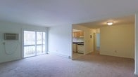 This two-floor space features an open-concept layout that seamlessly connects the main living area to the kitchen. The living room is finished with soft, neutral carpeting and bathed in natural light through a large picture window that looks out onto an outdoor view, creating a bright and airy atmosphere. A baseboard heater runs along the exterior wall beneath the window, providing efficient in-room climate control.

The kitchen is accessible directly from the living area through an open pass-through design, offering a functional and connected flow. It is equipped with wooden cabinetry, a gas range, and a dishwasher, with ample counter space and under-cabinet lighting that keeps the area well-lit. Adjacent to the kitchen, a hallway leads to additional rooms, suggesting a practical and well-organized floor plan across both levels.

The overall color palette is light and neutral throughout, with white walls and beige tones contributing to a clean, understated aesthetic. The space is practical in its layout, offering clear sightlines between living and cooking areas, making it suitable for everyday living.