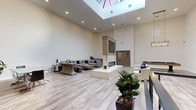 Fifty58 is a thoughtfully designed two-floor venue that blends contemporary aesthetics with versatile, functional spaces throughout. The main level immediately impresses with its soaring double-height ceilings and a large overhead skylight that floods the open floor plan with natural light. Pale wood-look flooring runs throughout, complementing the neutral, warm color palette. A central marble-clad fireplace with a mounted television above serves as the focal point of the main gathering area, surrounded by generously sized sectional sofas, lounge chairs, and accent furnishings arranged for both casual relaxation and social gatherings. An upper mezzanine level, accessible by a sleek staircase with modern cable railings, offers additional seating nooks accented by a striking sputnik chandelier. The lower level provides a more intimate atmosphere with recessed lighting and multiple distinct zones, including a large lounge area with oversized sectionals and a wall-mounted television, a dedicated game room with a poker-style table and upholstered chairs, a private meeting room with a glass-top conference table, and a bar counter with pendant lighting and barstool seating. A pool table rounds out the recreational offerings. Throughout both floors, curated greenery and tasteful artwork contribute to a polished, welcoming ambiance.