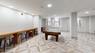 Located at 31 Pratt St in downtown Hartford, Connecticut, this amenity lounge is a well-appointed community gathering space designed for relaxation and recreation. The single-floor layout offers a generous open area unified by a striking geometric-patterned tile floor in warm neutral tones, which serves as a defining design element throughout the space.

At the heart of the room sits a full-size billiards table with a rich wood frame, positioned beneath recessed ceiling lighting that evenly illuminates the space. A long bar-height wooden counter with woven stools lines one wall, providing an ideal spot for casual socializing or spectating. Natural light filters in through multiple windows, offering views of the surrounding Hartford cityscape.

The lounge area features a sectional sofa arranged around a round coffee table, complemented by an accent chair and side table on a layered area rug. A wall-mounted flat-screen TV is positioned in a dedicated media nook, creating a comfortable viewing corner. Curated abstract artwork adorns the walls, adding a polished, contemporary aesthetic. The color palette blends crisp white ceilings with warm taupe accent walls, contributing to an inviting and modern ambiance. A black-framed entry door with sidelights completes the thoughtfully designed space.