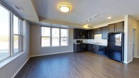 This single-floor space showcases a seamless open-concept layout that integrates the living area and kitchen into one cohesive environment. The rich, dark hardwood-style flooring runs throughout, providing a striking contrast against the light-toned walls and creating a sense of warmth and continuity across the space.

The kitchen is equipped with dark espresso cabinetry that extends to the upper and lower walls, offering generous storage. Black appliances, including a range, microwave, and refrigerator, complement the cabinet finish, while light-toned countertops provide a functional and visually balanced workspace. A double sink sits beneath under-cabinet lighting, adding both practicality and ambiance.

A large picture window in the living area serves as a standout feature, flooding the space with natural light while framing a view of the surrounding urban skyline. The open floor plan allows for flexible furniture arrangement in the living area, making the space feel expansive and versatile.

Ceiling-mounted flush lighting and a track lighting system ensure the space is well-lit throughout. The overall aesthetic is modern and contemporary, with a cohesive dark-toned palette balanced by lighter walls and ceilings.