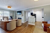 Located at 6075 Grelot Rd in Mobile, Alabama, this single-floor space offers a thoughtfully designed open-concept layout finished with warm hardwood-style flooring and soft sage-green walls throughout. Crown molding adds a refined architectural touch to each room, contributing to a cohesive and polished aesthetic.

The main living and office area is generously proportioned, centered around a classic fireplace with a white mantel and a wall-mounted flat-screen television above. Large windows allow natural light to fill the room, creating a bright and inviting atmosphere. A wood-toned desk and seating arrangement make this space equally suited for professional use.

The galley-style kitchen is efficiently laid out with crisp white shaker cabinetry, sleek brushed-nickel hardware, dark granite countertops, and stainless steel appliances including a refrigerator and microwave. Recessed lighting illuminates the workspace, and light wood-look flooring ties the kitchen seamlessly to the rest of the unit.

The entryway offers a welcoming first impression, featuring a charming wooden bench, a woven area rug, and a glass-paneled front door that draws in additional natural light. Decorative greenery accents the space, reinforcing the warm and approachable character found throughout this well-appointed Mobile property.