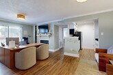 Located at 6075 Grelot Rd in Mobile, Alabama, this single-floor space offers a thoughtfully designed open-concept layout finished with warm hardwood-style flooring and soft sage-green walls throughout. Crown molding adds a refined architectural touch to each room, contributing to a cohesive and polished aesthetic.

The main living and office area is generously proportioned, centered around a classic fireplace with a white mantel and a wall-mounted flat-screen television above. Large windows allow natural light to fill the room, creating a bright and inviting atmosphere. A wood-toned desk and seating arrangement make this space equally suited for professional use.

The galley-style kitchen is efficiently laid out with crisp white shaker cabinetry, sleek brushed-nickel hardware, dark granite countertops, and stainless steel appliances including a refrigerator and microwave. Recessed lighting illuminates the workspace, and light wood-look flooring ties the kitchen seamlessly to the rest of the unit.

The entryway offers a welcoming first impression, featuring a charming wooden bench, a woven area rug, and a glass-paneled front door that draws in additional natural light. Decorative greenery accents the space, reinforcing the warm and approachable character found throughout this well-appointed Mobile property.