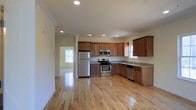 This single-floor residence offers a clean, functional layout with an emphasis on open living spaces and quality finishes throughout. The kitchen serves as a central highlight, featuring rich medium-toned wood cabinetry that provides ample storage, granite countertops, and a full suite of stainless steel appliances including a gas range with oven, built-in microwave, refrigerator, and dishwasher. A double-basin sink is positioned beneath a window, allowing for natural light while working in the kitchen. Recessed lighting is installed throughout the ceiling, ensuring well-distributed illumination across the space. The floors are finished in polished light hardwood, adding warmth and continuity as they extend seamlessly from the kitchen into the adjoining living areas. Crown molding accents the ceilings, lending a refined architectural detail to the interior. Multiple windows bring in generous natural light, contributing to an airy and welcoming ambiance. An open doorway visible from the kitchen connects to an adjacent room, reinforcing the home's open and connected floor plan. Overall, the space combines practical design with tasteful finishes, creating a comfortable and inviting living environment.