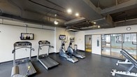This fitness facility offers a well-equipped, single-floor workout space designed to support a comprehensive exercise routine. The main area features multiple treadmills and elliptical machines arranged for easy access, each paired with individual wall-mounted monitors to keep users entertained during cardio sessions. Additional wall-mounted screens are positioned throughout the room for optimal viewing from any equipment.

The space showcases an industrial aesthetic, with exposed ceiling infrastructure including ductwork, pipes, and recessed and pendant lighting that illuminate the dark rubberized flooring. The clean white walls contrast with the utilitarian ceiling, creating a modern and functional atmosphere.

A glass-paneled door leads to an adjacent room, visible in the background, which appears to offer additional fitness equipment and further extends the workout capabilities of the facility. The open layout and thoughtful equipment placement ensure that the space remains practical and easy to navigate, making it suitable for a range of fitness activities. Overall, the facility combines a contemporary industrial design with essential amenities to deliver a focused and efficient exercise environment.