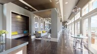 This contemporary single-floor space is designed with a refined, modern aesthetic that blends elegance with functionality. The interior is anchored by a striking reception counter finished with a marble-paneled front, wood trim, and a dark stone top, illuminated by a cluster of industrial-style pendant lights overhead. Soaring ceilings with dark coffered detailing and clerestory windows allow natural light to filter throughout the open-plan layout.

The main lounge area features plush gray seating accented with bold yellow and teal throw pillows, centered around a textured area rug and a weathered wood console. Decorative wall molding frames curated artwork, adding architectural character. A media nook with patterned wallcovering and wall-mounted televisions provides a casual gathering spot with high-top tables and sleek bar stools.

Along one side, floor-to-ceiling glass doors and windows flood the space with daylight and connect the interior to the outdoors. The polished large-format dark tile flooring runs throughout, adding a sleek, cohesive foundation.

Accessible from the interior, an outdoor rooftop terrace features a stone-clad built-in bar with a granite countertop, bar seating, and a pergola overhead with string lighting, offering panoramic open-sky views.