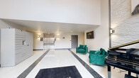 This generously sized single-floor common area serves as the central hub of the building, encompassing a grand entrance lobby, a resident lounge, a full kitchenette, and a restroom. The entrance lobby makes an immediate impression with soaring double-height ceilings, floor-to-ceiling windows that flood the space with natural light, and polished white tile flooring accented by a mosaic dark inlay strip. A branded welcome mat greets visitors at the glass entry doors, while smart parcel lockers, stainless steel elevator doors, and wall sconces contribute to a sleek, well-appointed atmosphere. Emerald green velvet seating adds a bold pop of color against the neutral walls. The resident lounge features wood-look plank flooring, a drop ceiling with recessed lighting, a round wooden dining table with upholstered chairs, and a striking series of geometric wall art panels. Lush potted plants enhance the welcoming ambiance. The kitchenette is equipped with white shaker cabinetry, quartz countertops, black hardware, stainless steel appliances, and a snack and beverage vending station. A neatly appointed restroom with a wood-front vanity and decorative wall planters completes the amenity suite.