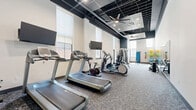 This fitness center offers a comprehensive workout environment designed to accommodate a wide range of exercise needs. The space features an open, elongated floor plan with generously high ceilings that lend an airy, expansive feel throughout. An exposed black ceiling with integrated ceiling fans, recessed lighting, and HVAC components contributes to the room's modern industrial aesthetic, while crisp white walls and patterned carpet flooring create a clean, energetic atmosphere.

Natural light filters in through multiple large windows fitted with roller shades along the main wall, providing a bright and welcoming environment. Wall-mounted flat-screen televisions are positioned at strategic intervals to keep users entertained during cardio sessions.

The cardio zone is well-appointed with multiple treadmills, elliptical trainers, and a stationary cycle. A dedicated strength training area features a multi-function cable machine and weight bench, catering to resistance and functional fitness needs. A distinct stretch and accessories corner at the far end of the room is outfitted with a rack of stability balls, resistance accessories, and a workout poster for guided exercises.

A large wall mirror enhances the sense of depth within the space, and a clearly marked exit door ensures safe and convenient access. Overall, the fitness center is thoughtfully organized and fully equipped to support both cardio and strength-focused training routines.